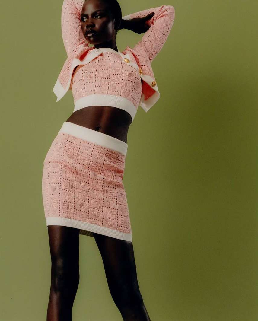 Adult female fashion model poses in a pink knit crop top, jacket, and skirt outfit against a green backdrop in a studio setting.