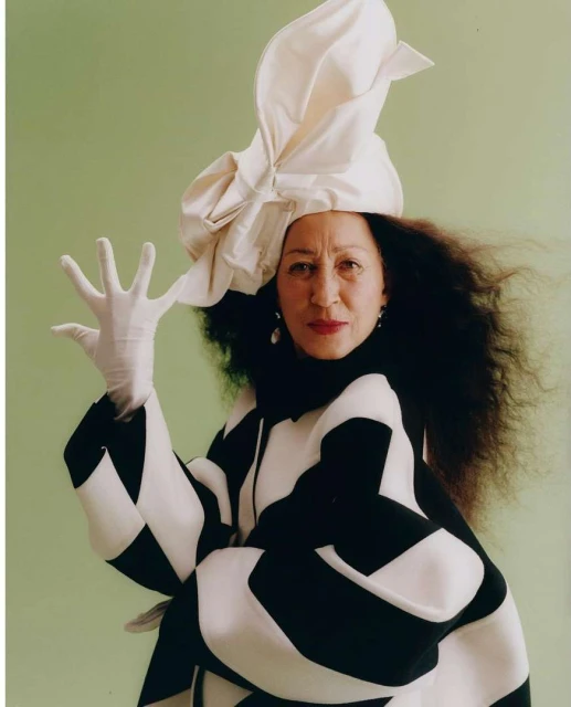 Portrait of an adult woman in a black and white checkered coat and a white sculptural hat, captured in a studio with a green background.