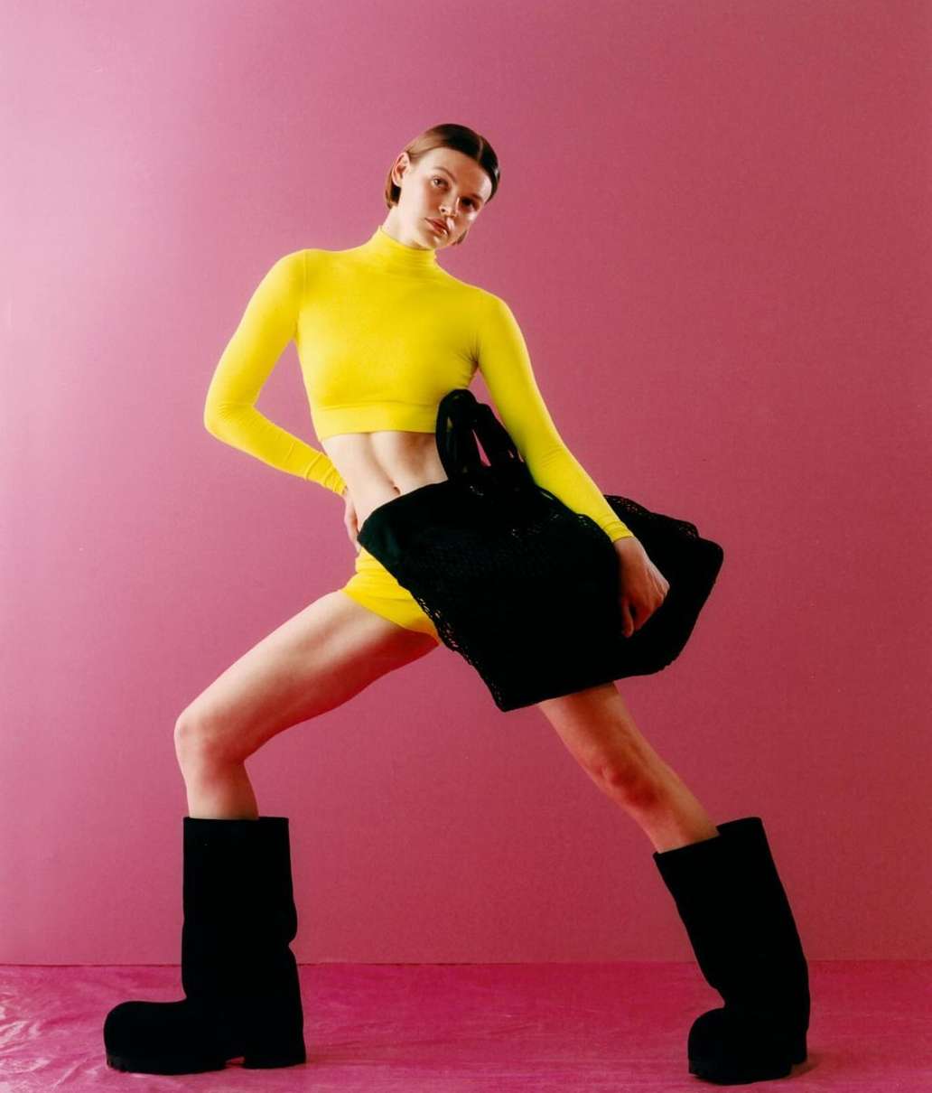 A fair-skinned woman strikes a pose in a yellow top and shorts, complemented by chunky black boots, against a vibrant pink backdrop.
