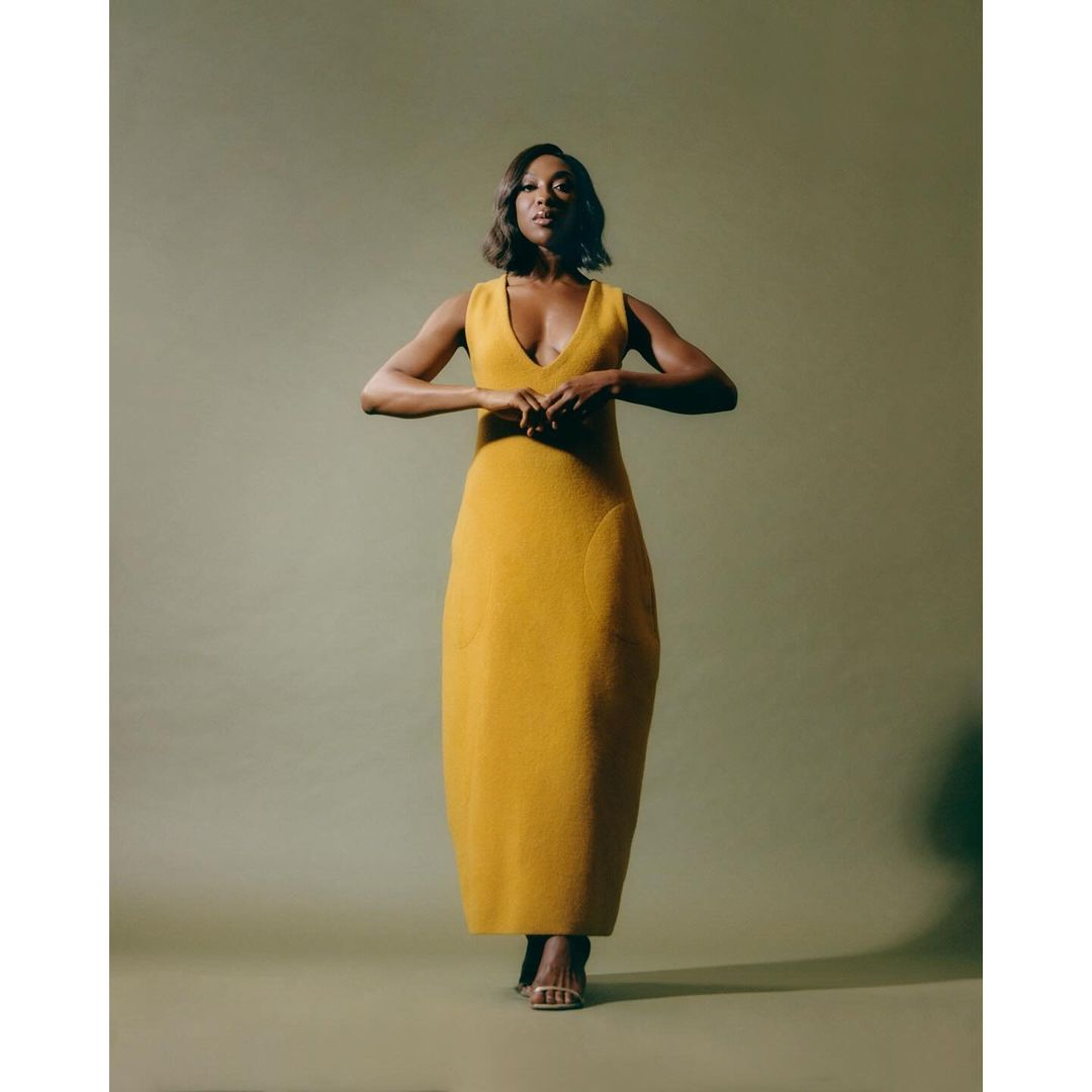 Elegant woman in a mustard yellow V-neck dress stands confidently with her hands gently clasped in front in a studio setting.