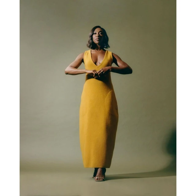 Elegant woman in a mustard yellow V-neck dress stands confidently with her hands gently clasped in front in a studio setting.