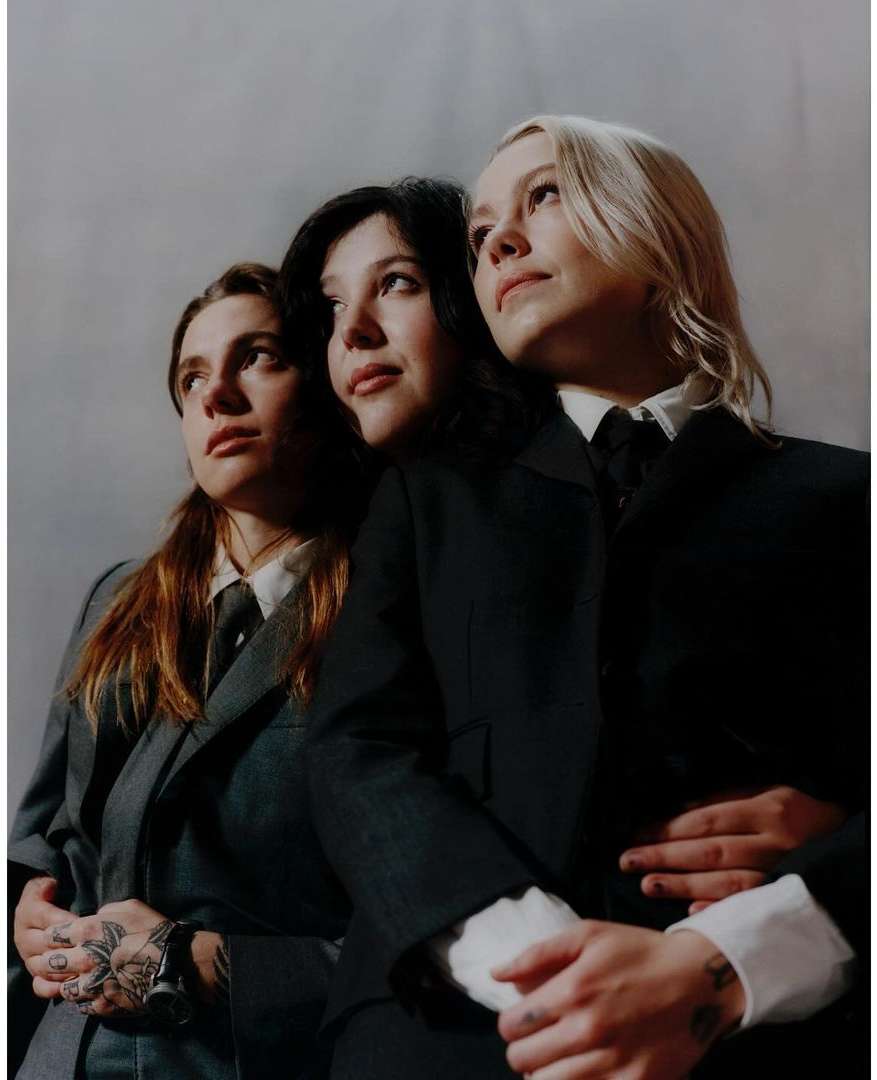 Three women in suits stand indoors against a grey backdrop. They face upwards, with two women holding hands and tattoo art visible.
