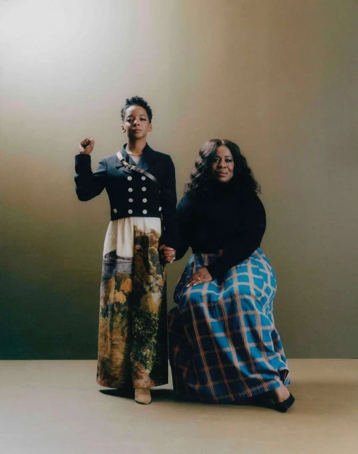 Two women stand indoors wearing long dresses; one raises a fist in the air. Soft, diffused lighting. Brown and beige color palette.