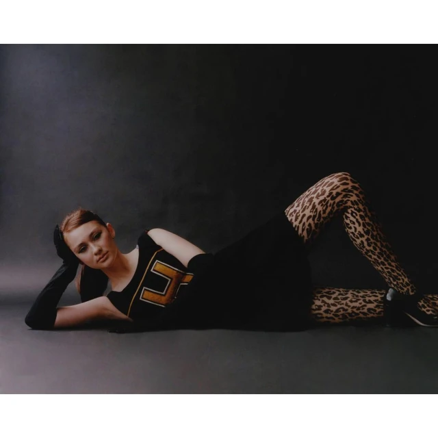 A woman in a black dress and leopard print tights reclines against a dark grey background, in a fashion studio setting with soft lighting.