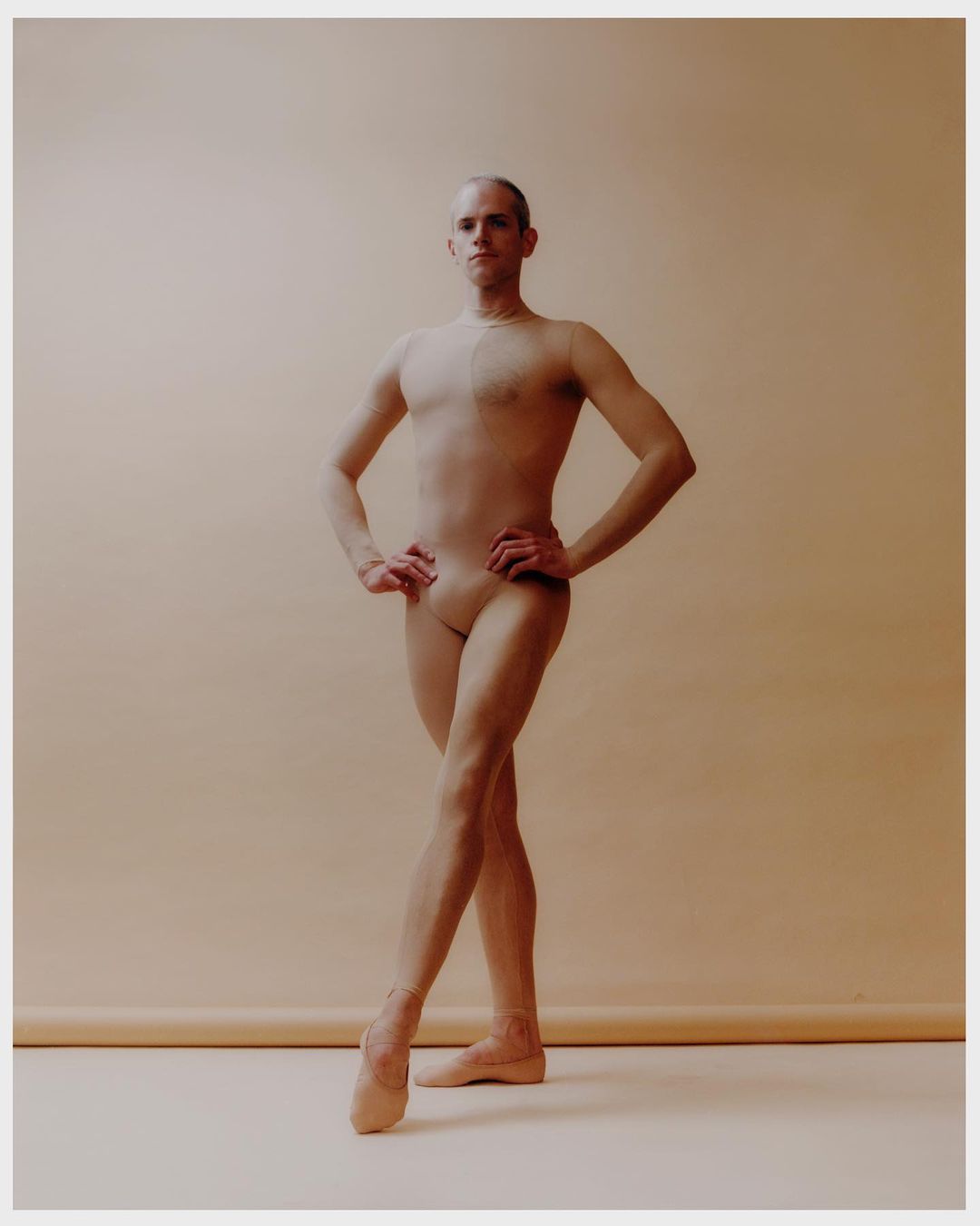 Full shot of a ballet dancer with short, light-brown hair posing with crossed legs in front of a neutral background in a dance studio.