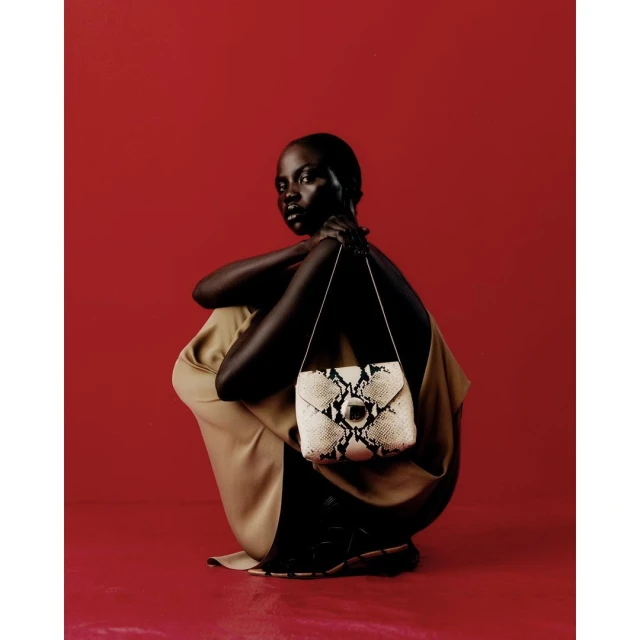 A dark-skinned woman in a crouched pose against a solid red background holds a white snakeskin purse with silver hardware.