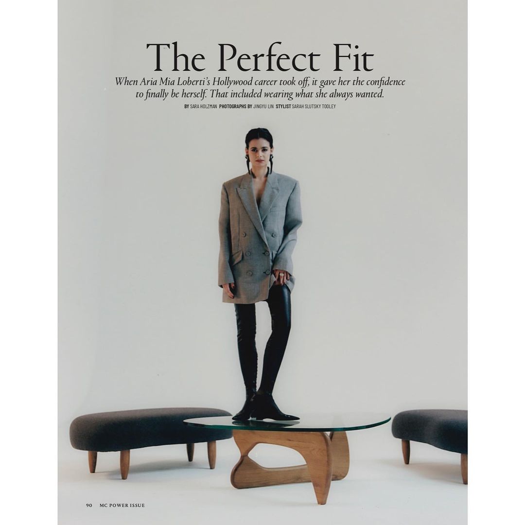 Fashion editorial shot of a young woman in an oversized blazer and black leggings, standing on a glass coffee table in a studio.