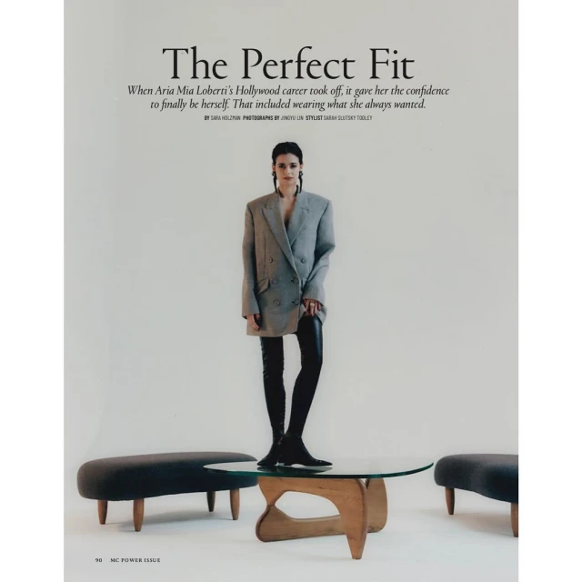 Fashion editorial shot of a young woman in an oversized blazer and black leggings, standing on a glass coffee table in a studio.