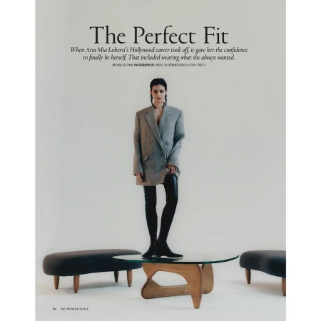 Fashion editorial shot of a young woman in an oversized blazer and black leggings, standing on a glass coffee table in a studio.