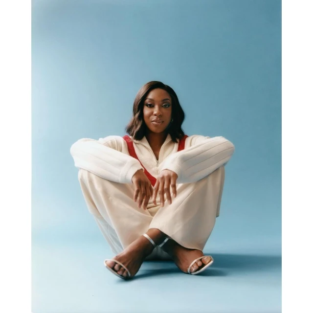 Stylish woman in a white jumpsuit with red straps and strappy heels sits cross-legged against a light blue backdrop in a studio shot.