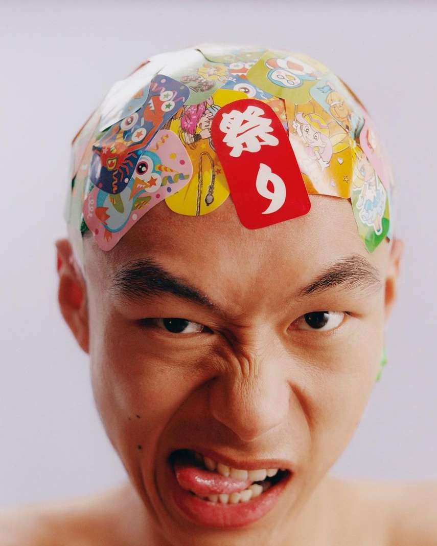 Close-up studio shot of an adult Asian male with a bare head covered in colorful stickers, sticking out his tongue and making a grimace.