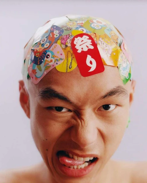 Close-up studio shot of an adult Asian male with a bare head covered in colorful stickers, sticking out his tongue and making a grimace.