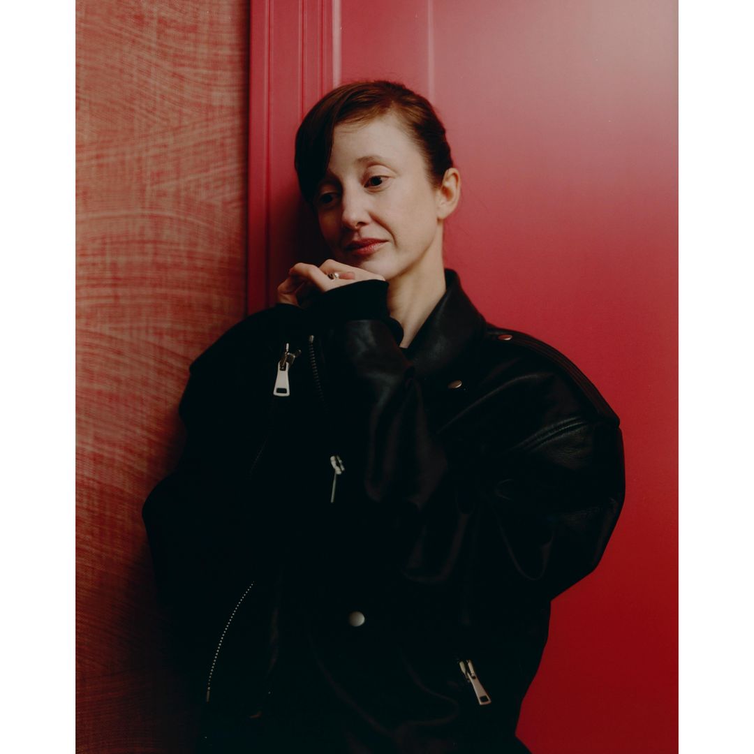 Portrait of a fair-skinned woman with auburn hair wearing a black leather jacket leaning against a red wall in an indoor setting.