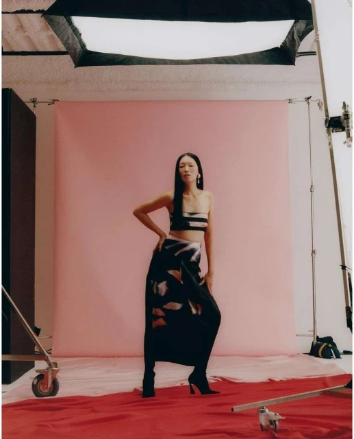 Asian woman poses in a two-piece outfit with a dark patterned skirt in a photo studio with pink backdrop and professional lighting.