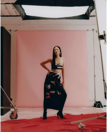 Asian woman poses in a two-piece outfit with a dark patterned skirt in a photo studio with pink backdrop and professional lighting.
