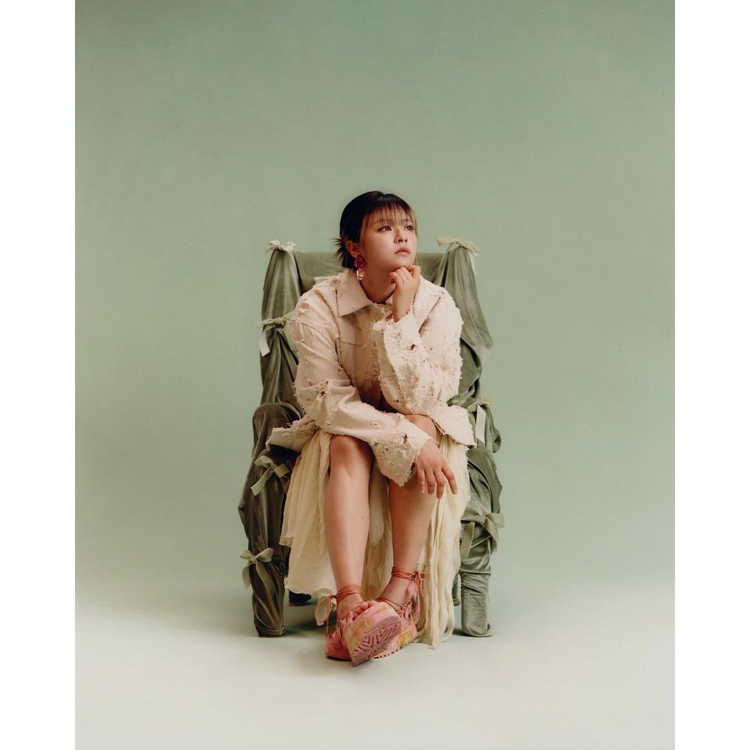 An Asian woman sits in an ornately decorated chair against a muted green backdrop, wearing a textured jacket, pink shoes and dangling earrings.