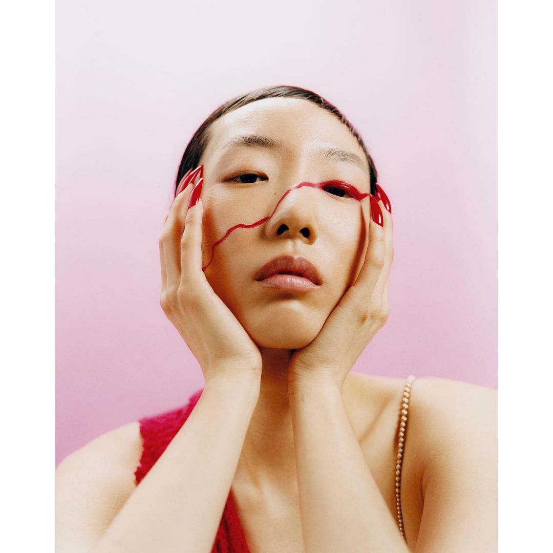 Asian woman portrait with hands on face, red nails, red line detail on face, against gradient pink background, beauty shot.