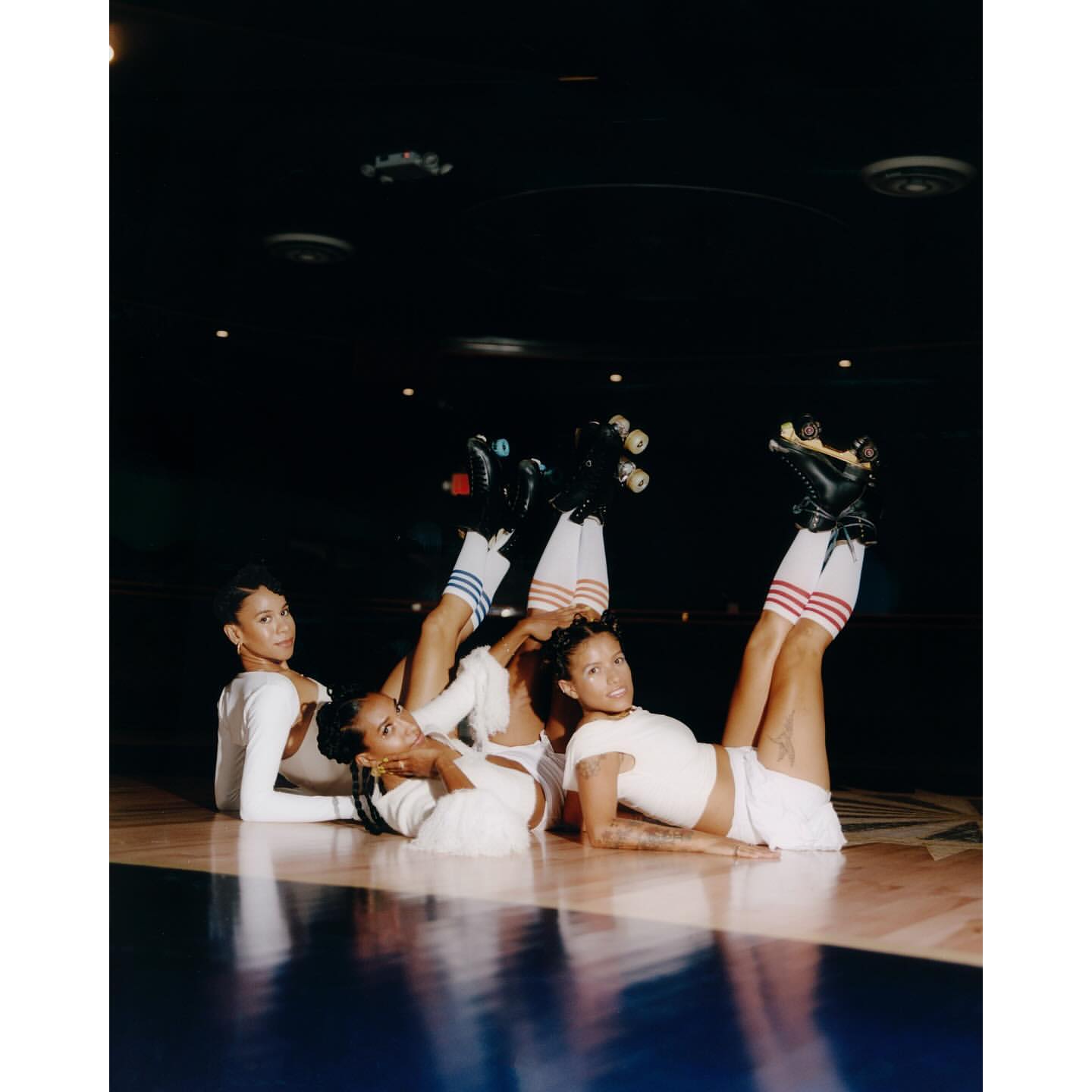Three women strike a pose indoors, lying on the floor wearing roller skates, striped socks and white tops, with a dark background.