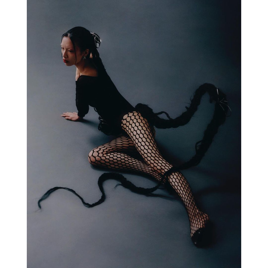 A woman wearing a black dress and fishnet stockings poses on a gray surface, with her long braids dramatically framing her body.