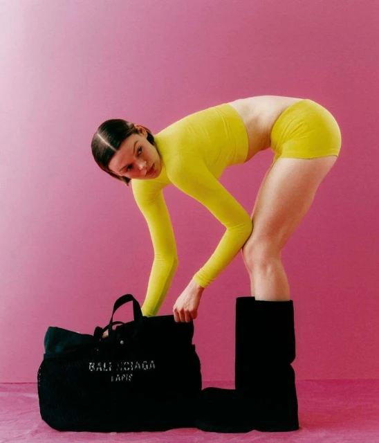 Fashion shot of a woman in a yellow outfit leaning over a black Balenciaga bag, set against a vibrant pink backdrop in a photo studio.