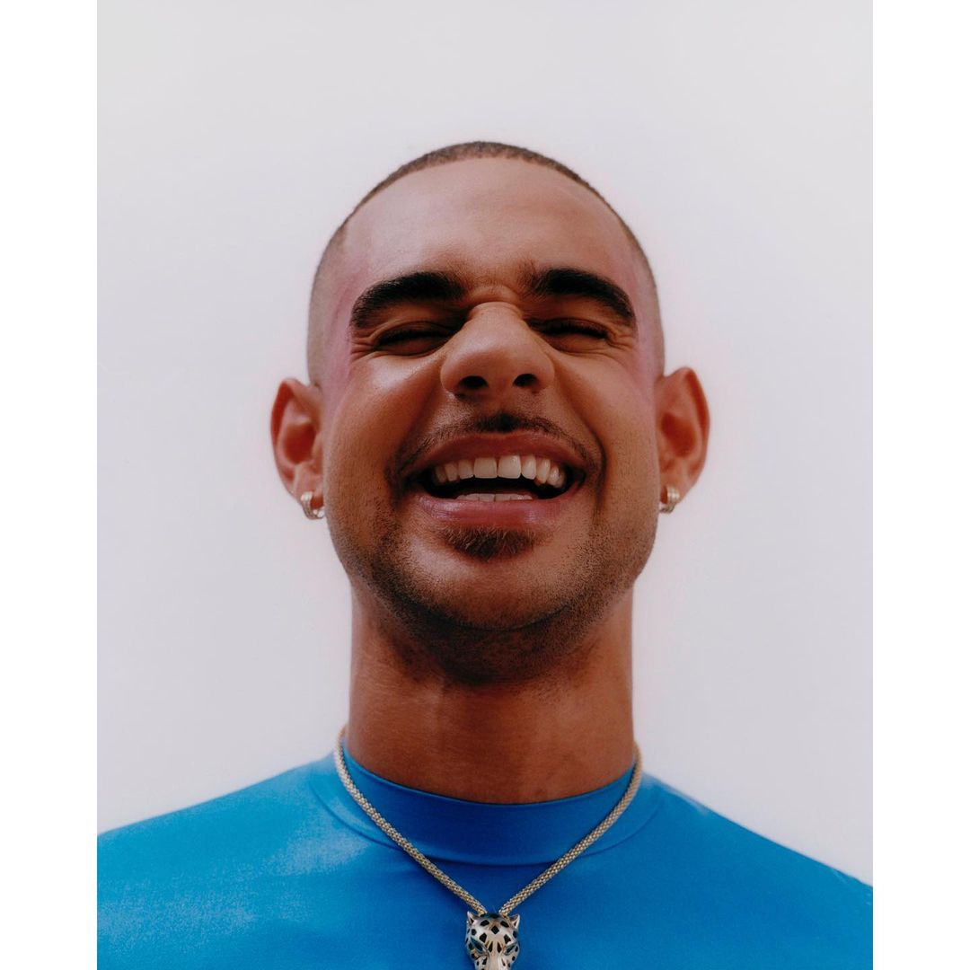 Close-up portrait of a smiling man wearing a blue top and a silver animal pendant necklace against a light background.