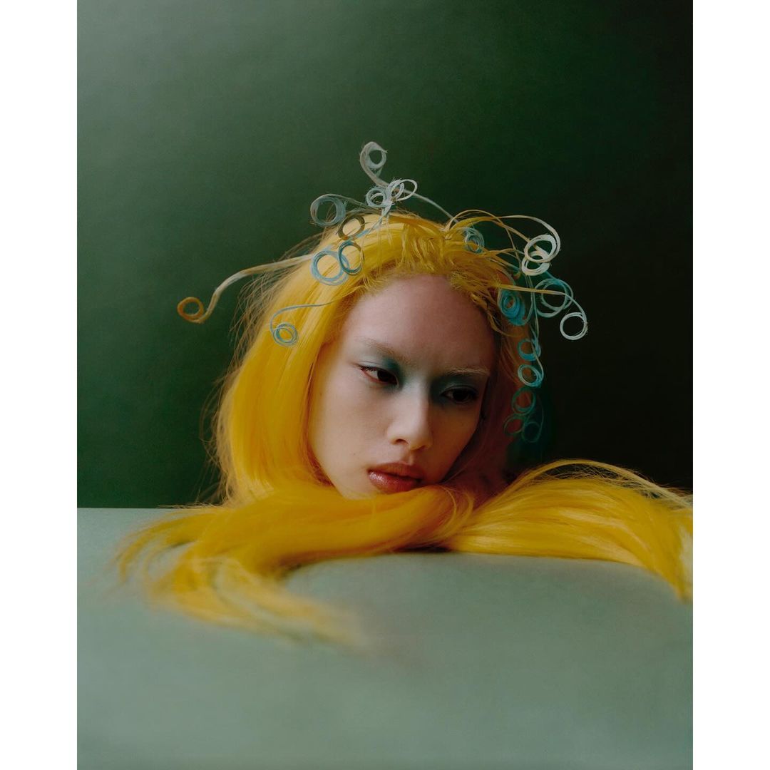 Surreal portrait of a woman with yellow hair and blue eye makeup with artistic headpiece, against a dark green backdrop indoors.