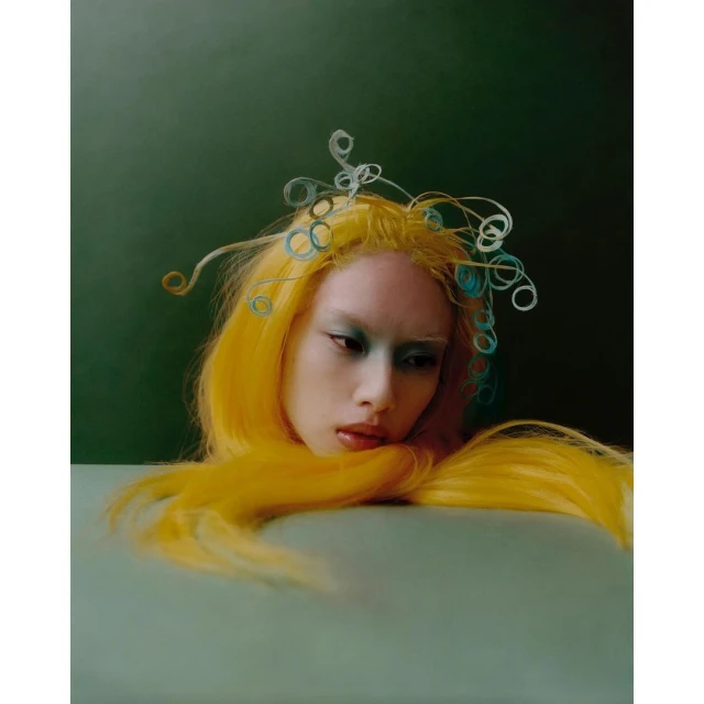 Surreal portrait of a woman with yellow hair and blue eye makeup with artistic headpiece, against a dark green backdrop indoors.
