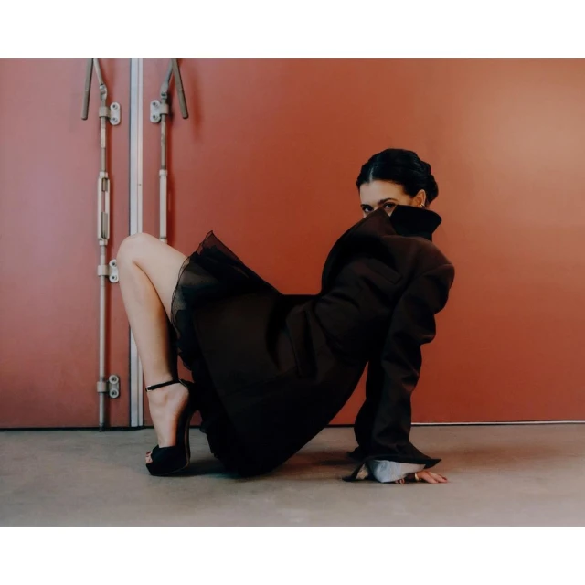 Adult female fashion model poses in a dramatic black coat with a short skirt in front of a salmon-colored wall and double doors.