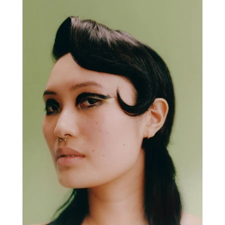 Close-up portrait of a woman with dark styled hair, bold eyeliner, and a septum piercing against a green background