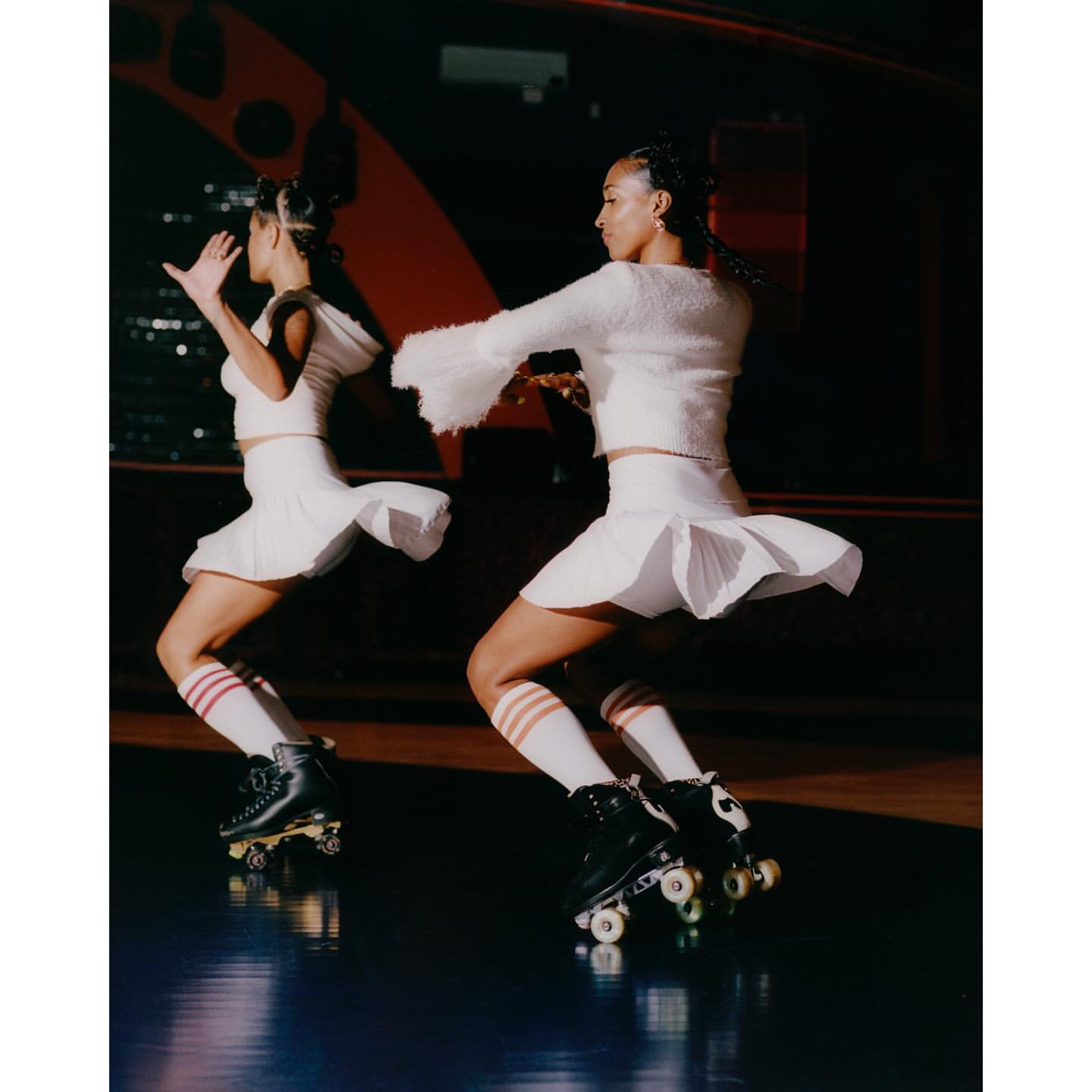 Two adult females skating in a roller rink wearing white pleated skirts, sweaters, striped socks, and black roller skates.