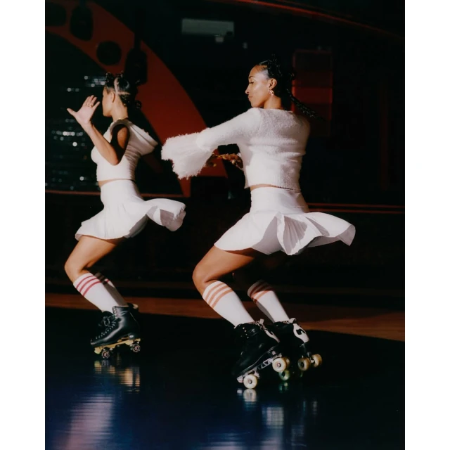Two adult females skating in a roller rink wearing white pleated skirts, sweaters, striped socks, and black roller skates.