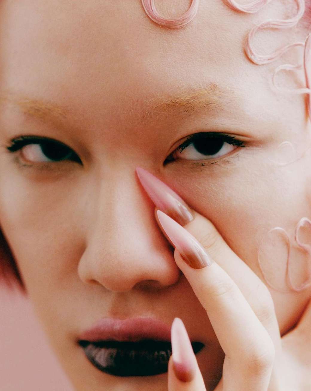 Close-up portrait of a woman with pale skin, pink decorations in her hair, dark glossy lips, and long, pink-ombre stiletto nails.