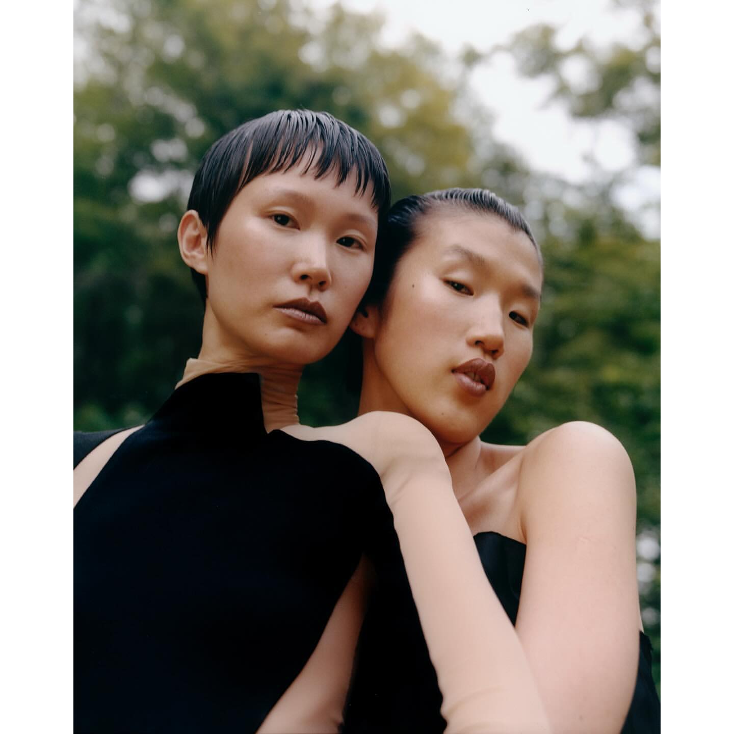 Two young adult female models in black strapless dresses pose closely together outdoors against a blurred green leafy backdrop in a fashion editorial.