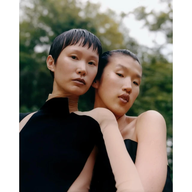 Two young adult female models in black strapless dresses pose closely together outdoors against a blurred green leafy backdrop in a fashion editorial.