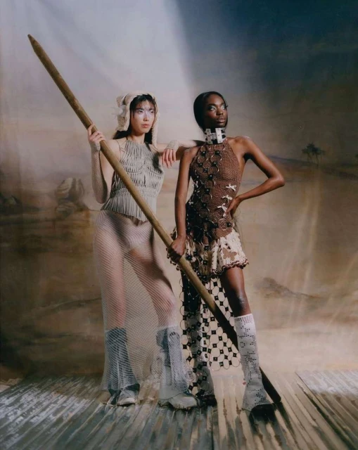 Two fashion models in conceptual clothing pose indoors against a painted desert backdrop, holding a tall pole. The lighting is soft and diffused.