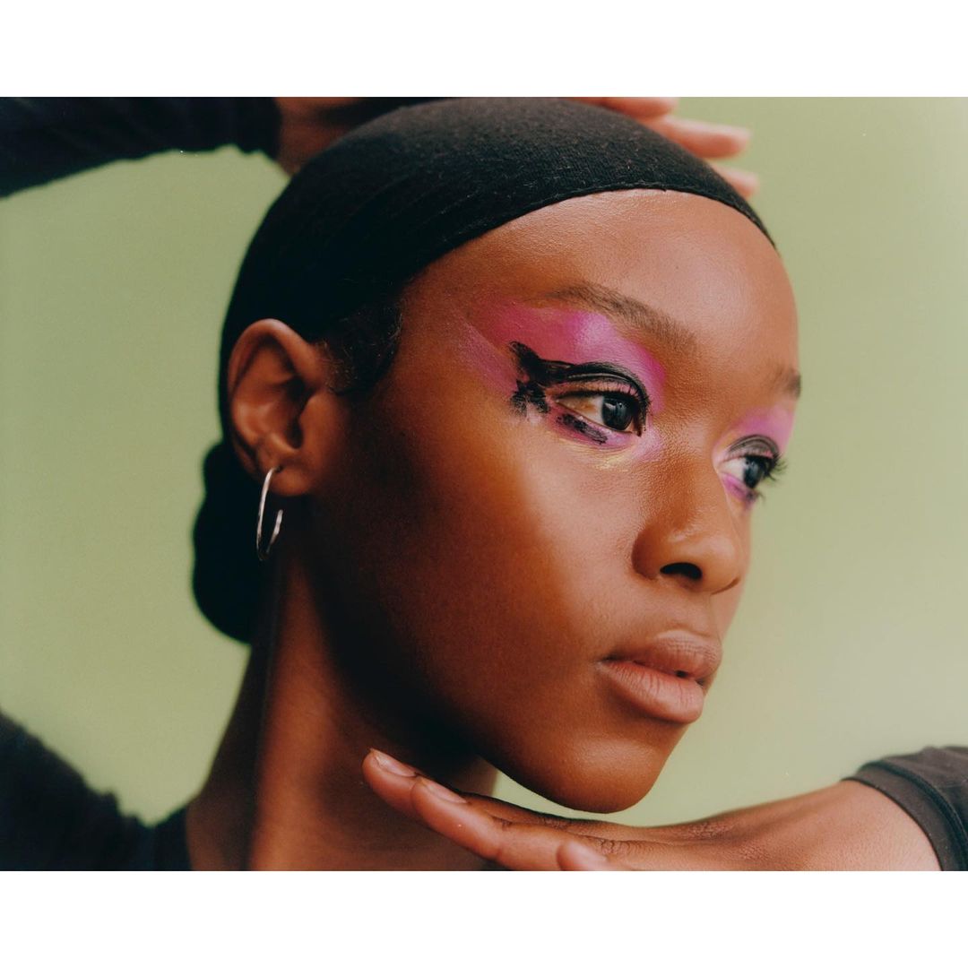 Close-up of a young African-American woman with creative pink and black eye makeup and silver hoop earrings against a light green background.