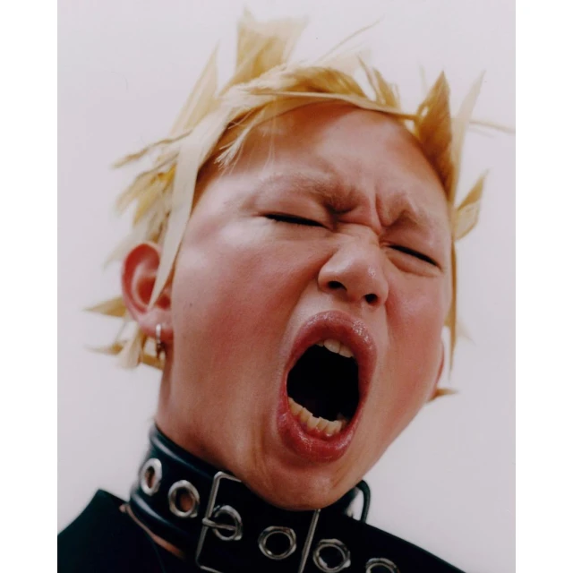 Portrait of a young adult with light-colored spiky hair, an expressive face with eyes closed, and a black studded collar necklace.