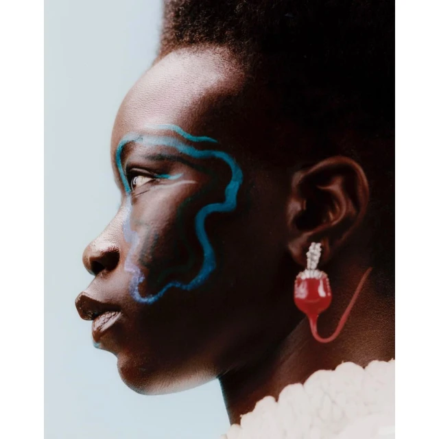 Close-up of a dark-skinned woman with artistic blue makeup, wearing a large jeweled red earring, against a solid light-blue background.