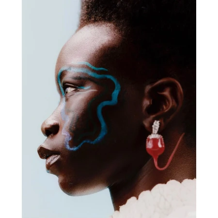 Close-up of a dark-skinned woman with artistic blue makeup, wearing a large jeweled red earring, against a solid light-blue background.