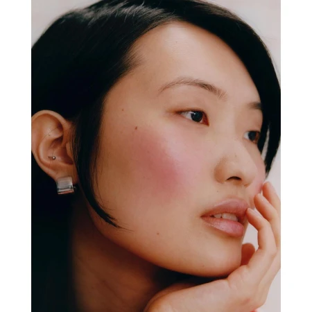 Close-up portrait of an East Asian woman with dark hair, a silver earring, and fingers resting near her chin against a neutral background.