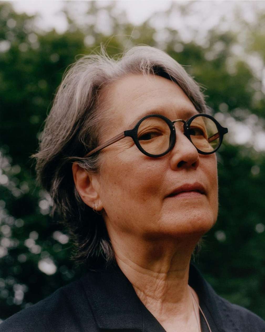 Close-up portrait of an adult woman with short grey hair and round glasses looking upwards in a green outdoor setting.