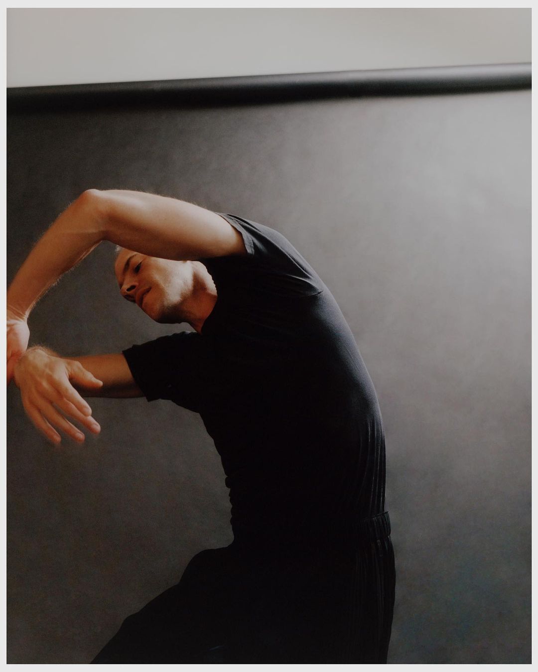 Male dancer in black activewear poses gracefully against a dark background, showcasing flexibility and artistry through dynamic movement.