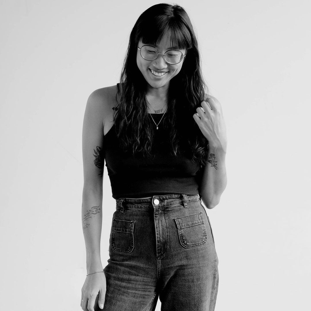 Monochrome portrait of a smiling woman with glasses wearing a black tank top and denim jeans against a plain backdrop.
