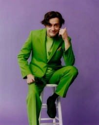 Man wearing a bright green suit and matching t-shirt seated on a white stool against a solid purple backdrop.
