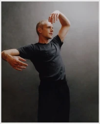 A man in a dark grey t-shirt posing with graceful hand gestures against a textured grey studio background.