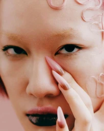 Close-up portrait of a model with sculpted pink hair details on skin, dark eyeliner, dark lips, and long pointed pink nails.