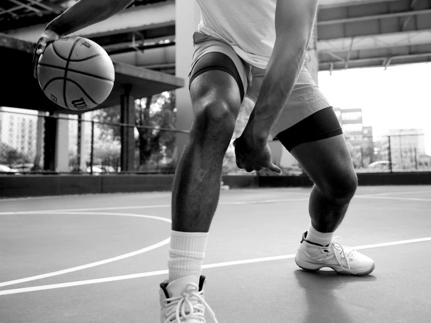 Monochrome close-up of a muscular adult male athlete dribbling a Wilson basketball on a public court outdoors in daylight.