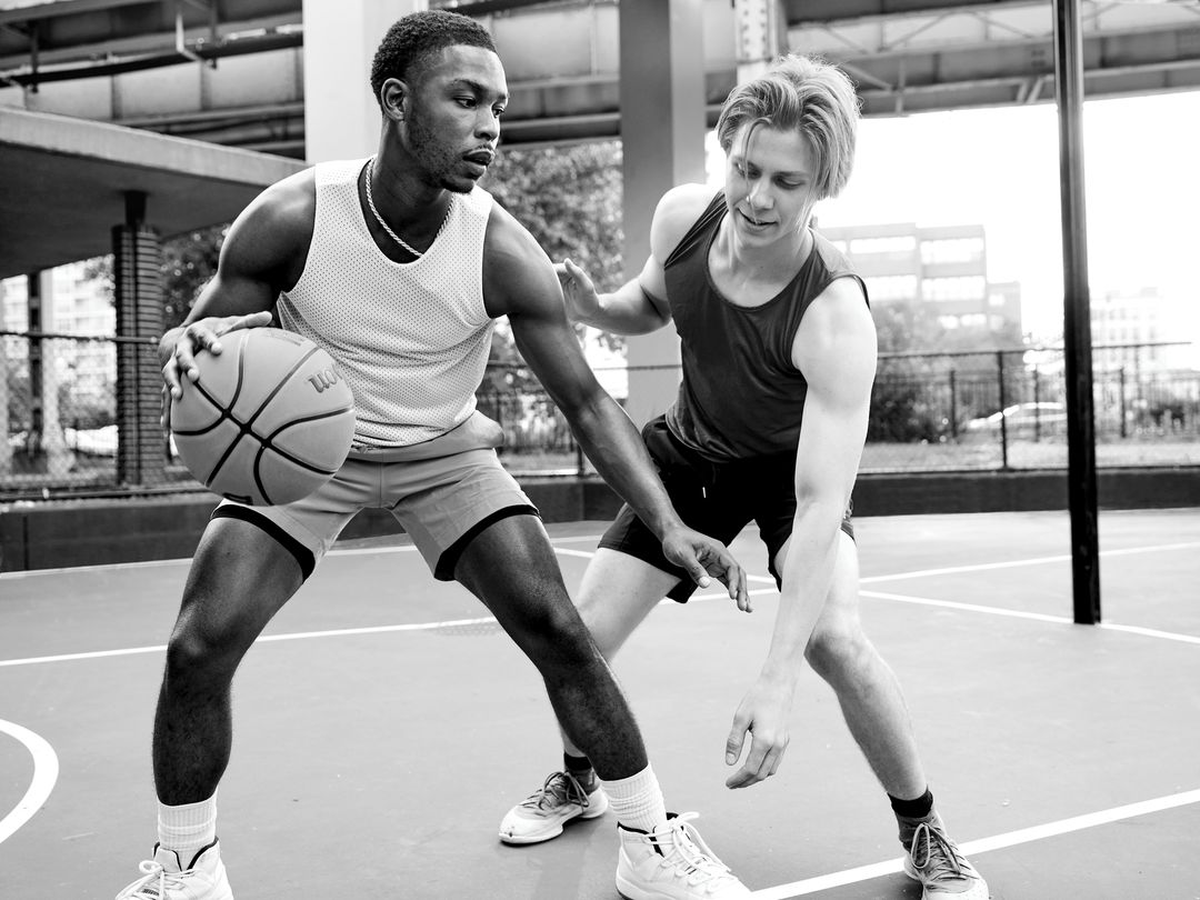 Two athletic men play basketball outdoors on a grey court. One dribbles the ball, wearing a white tank top and grey shorts.