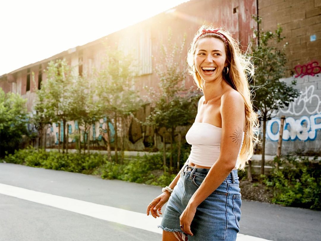 Happy woman with a headband in denim shorts, white tube top, and hoop earrings laughs outdoors next to a building with graffiti.