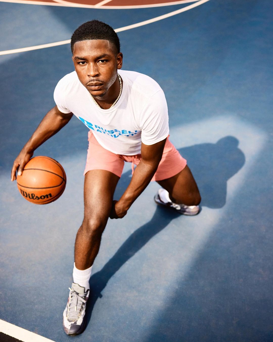 Athletic Black male dribbling a Wilson basketball on a blue sports court in sunlight, wearing shorts, t-shirt, and sneakers.